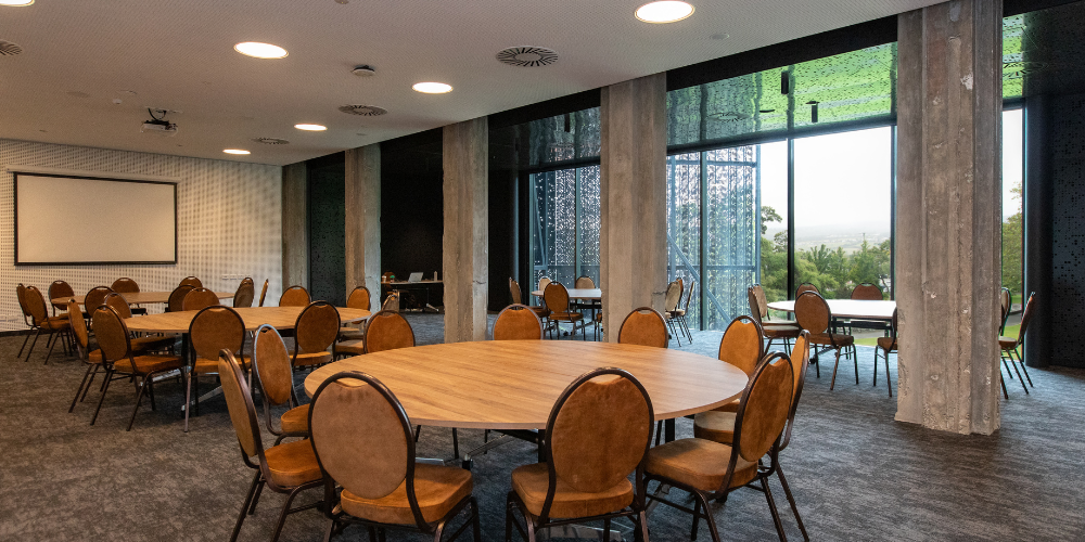 The Tamar Valley Room set for a banquet, with round tables, upholstered chairs, a presentation screen and views across the landscape through the glass windows.